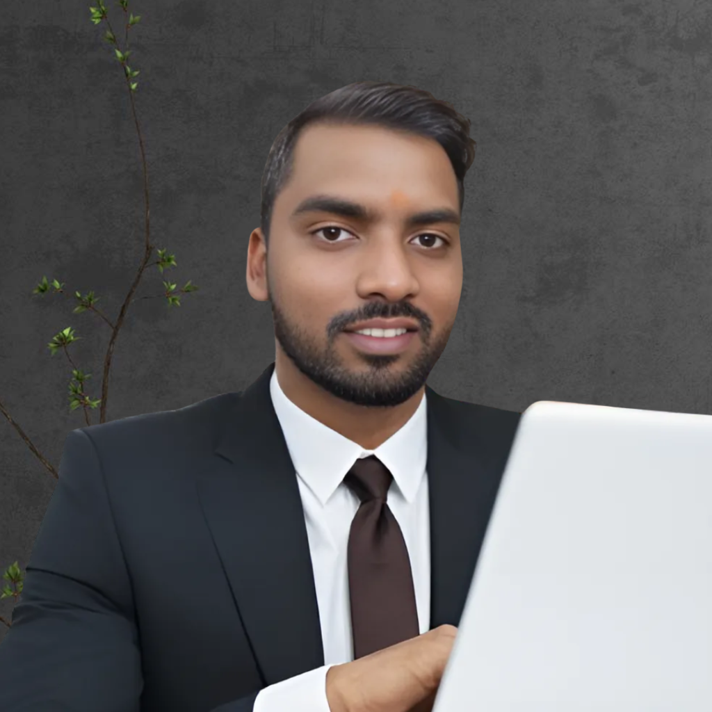 A professionally dressed man in a black suit, white shirt, and brown tie, working on a laptop with a confident smile. The background features a dark textured wall with a small green plant.
